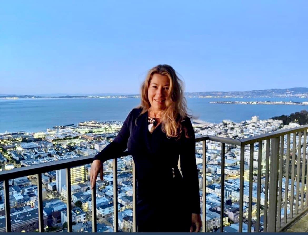 Milena Gross on a balcony overlooking the San Francisco skyline and bay at dusk.
