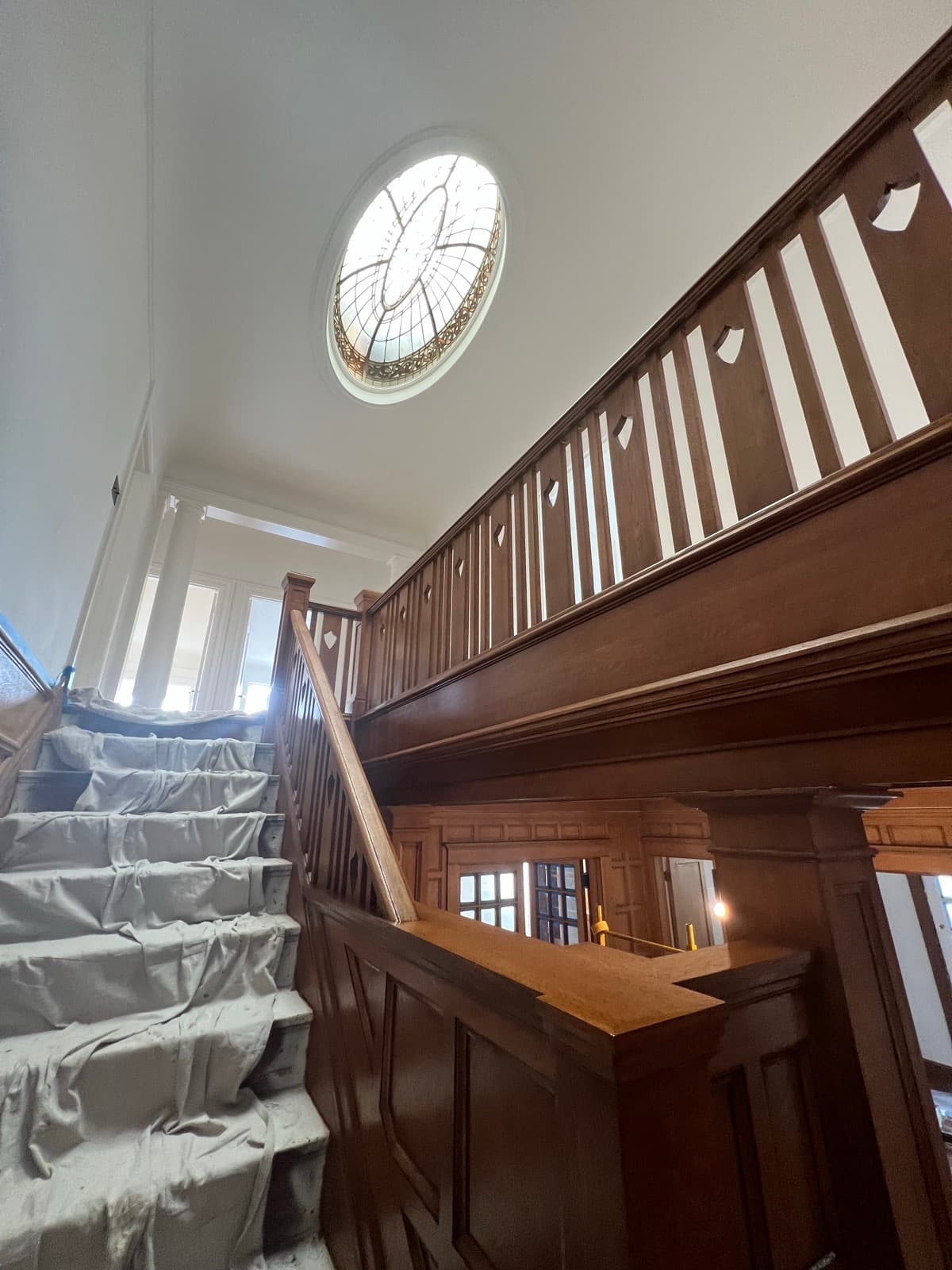 The staircase part-way through: the railing and paneling already stripped back to wood, the treads still under drop cloths, the oval leaded-glass skylight overhead